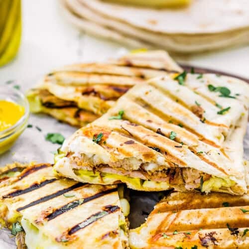 Easy Cuban Quesadillas: Grilled quesadillas filled with shredded meat, melted cheese, and pickles, garnished with chopped herbs and served on a wooden board with tortillas and cheese in the background.