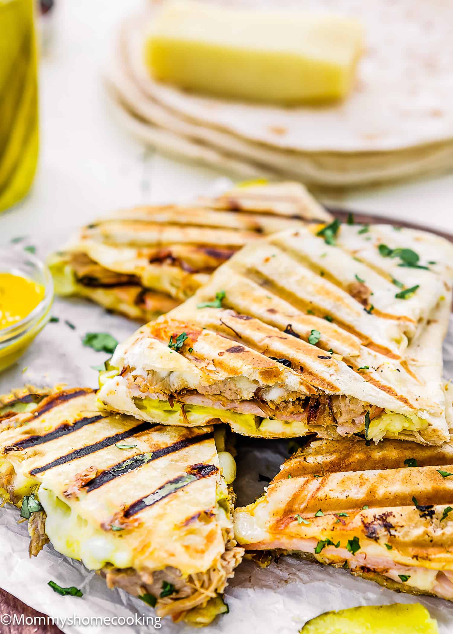 Easy Cuban Quesadillas: Grilled quesadillas filled with shredded meat, melted cheese, and pickles, garnished with chopped herbs and served on a wooden board with tortillas and cheese in the background.
