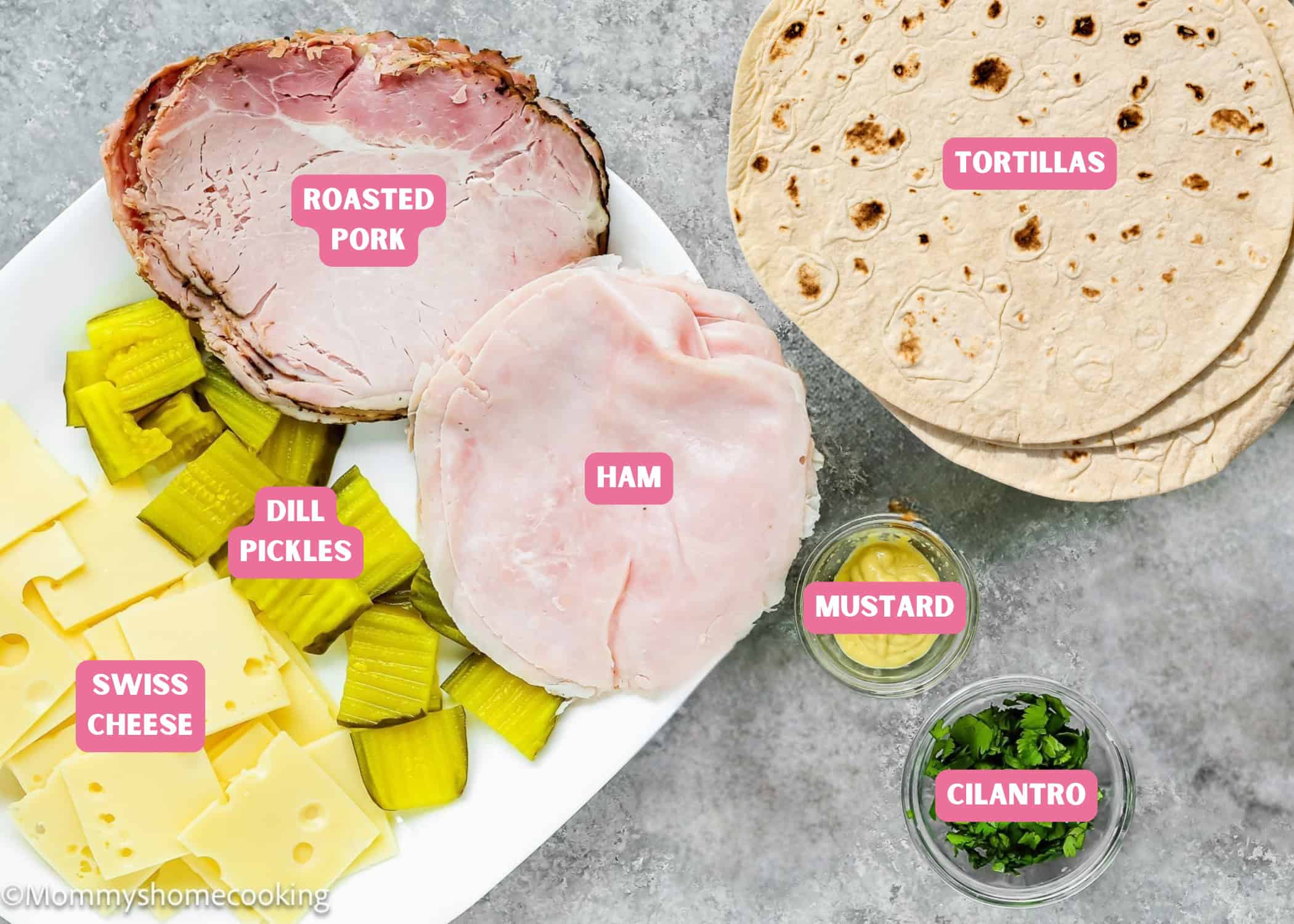 Plate with roasted pork, ham, Swiss cheese, and dill pickles alongside tortillas, small bowls of mustard, and chopped cilantro on a gray surface. Perfect for making Easy Cuban Quesadillas. Each item is labeled.