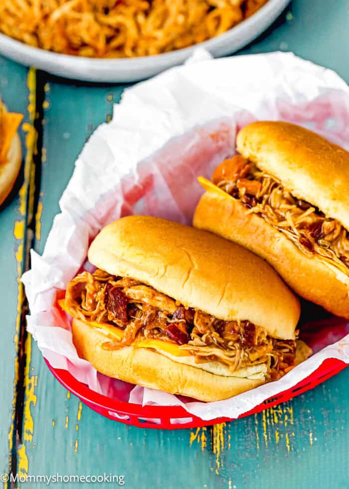 Easy Pulled Barbecue Chicken Sandwiches with barbecue sauce are served in a red basket lined with white parchment paper, with extra shredded chicken in a dish in the background.