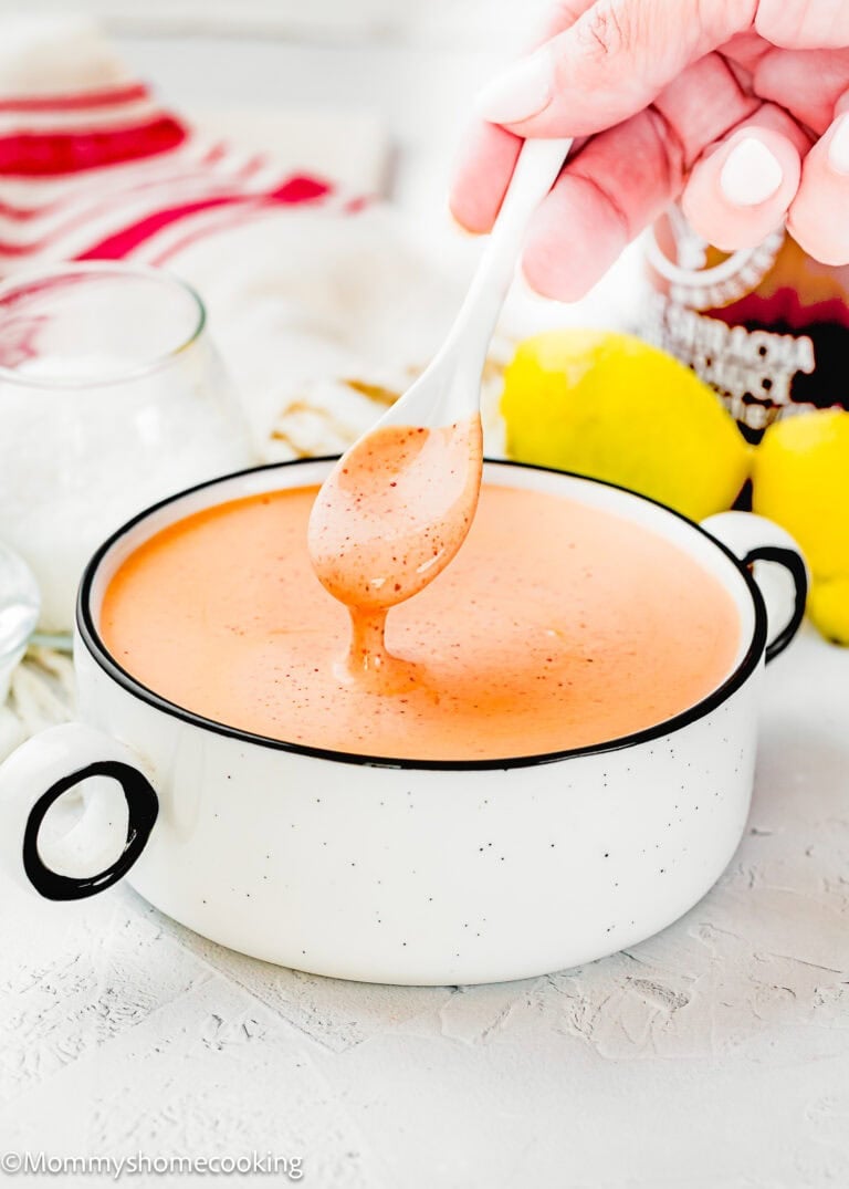 A hand holds a white spoon above a white bowl filled with creamy, orange Easy Spicy Mayo Sauce. Lemons and a glass jar are in the background.