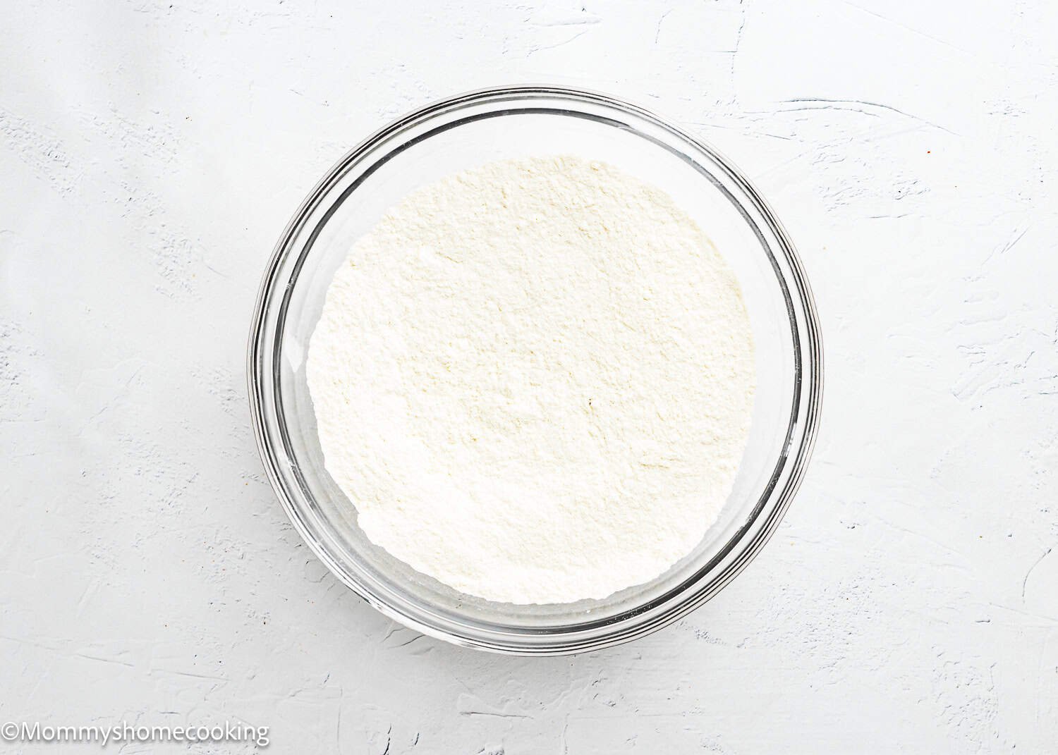 A clear glass bowl filled with white flour sits on a white textured surface, ready for making Eggless Chocolate Chip Cookie Bars.