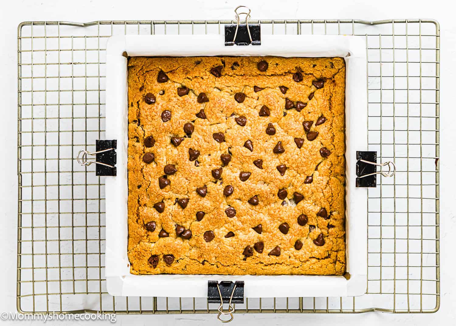 An eggless chocolate chip cookie bar in a parchment-lined baking pan, secured with binder clips, sits on a cooling rack.