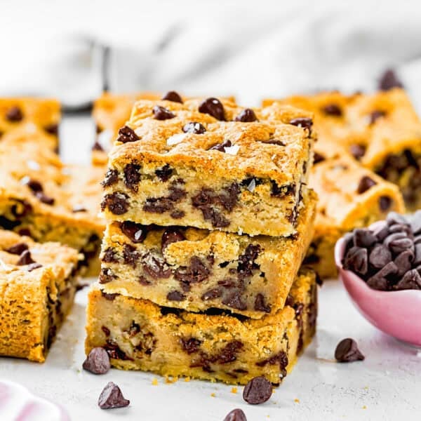 Three stacked Eggless Chocolate Chip Cookie Bars are in focus, with more bars and a small bowl of chocolate chips in the background.
