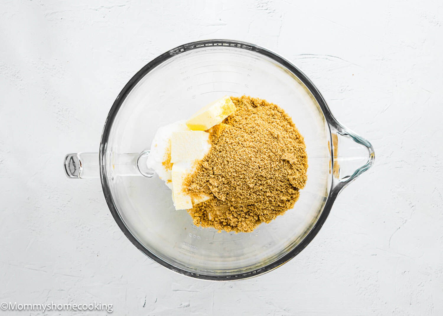 A glass mixing bowl containing granulated sugar, brown sugar, and chunks of butter on a white surface for making Eggless Chocolate Chip Cookie Bars.