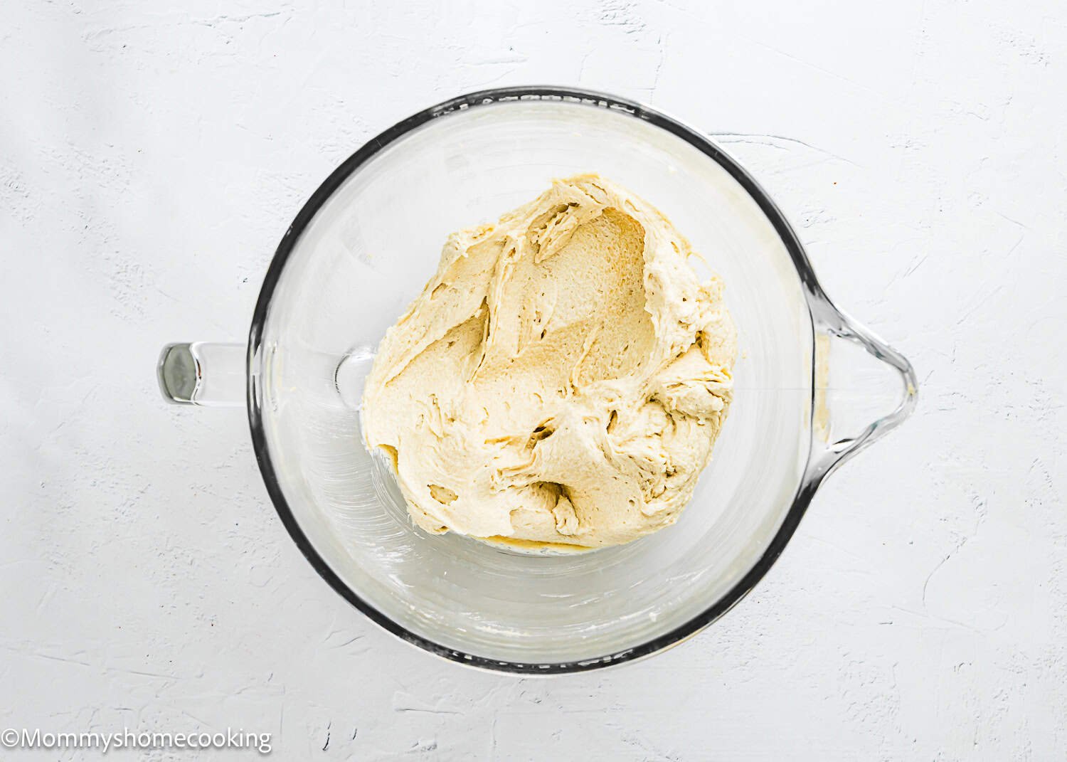 A glass mixing bowl containing a portion of creamy, pale-colored dough for Eggless Chocolate Chip Cookie Bars sits on a white surface.