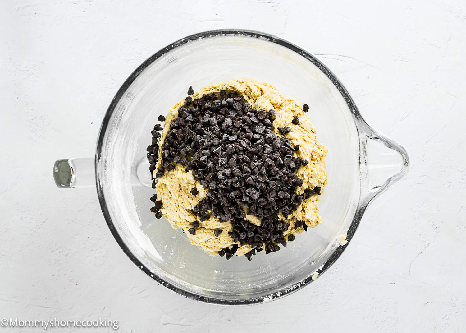 A glass mixing bowl with partially mixed Eggless Chocolate Chip Cookie Bars dough and a pile of chocolate chips on top, viewed from above on a white surface.
