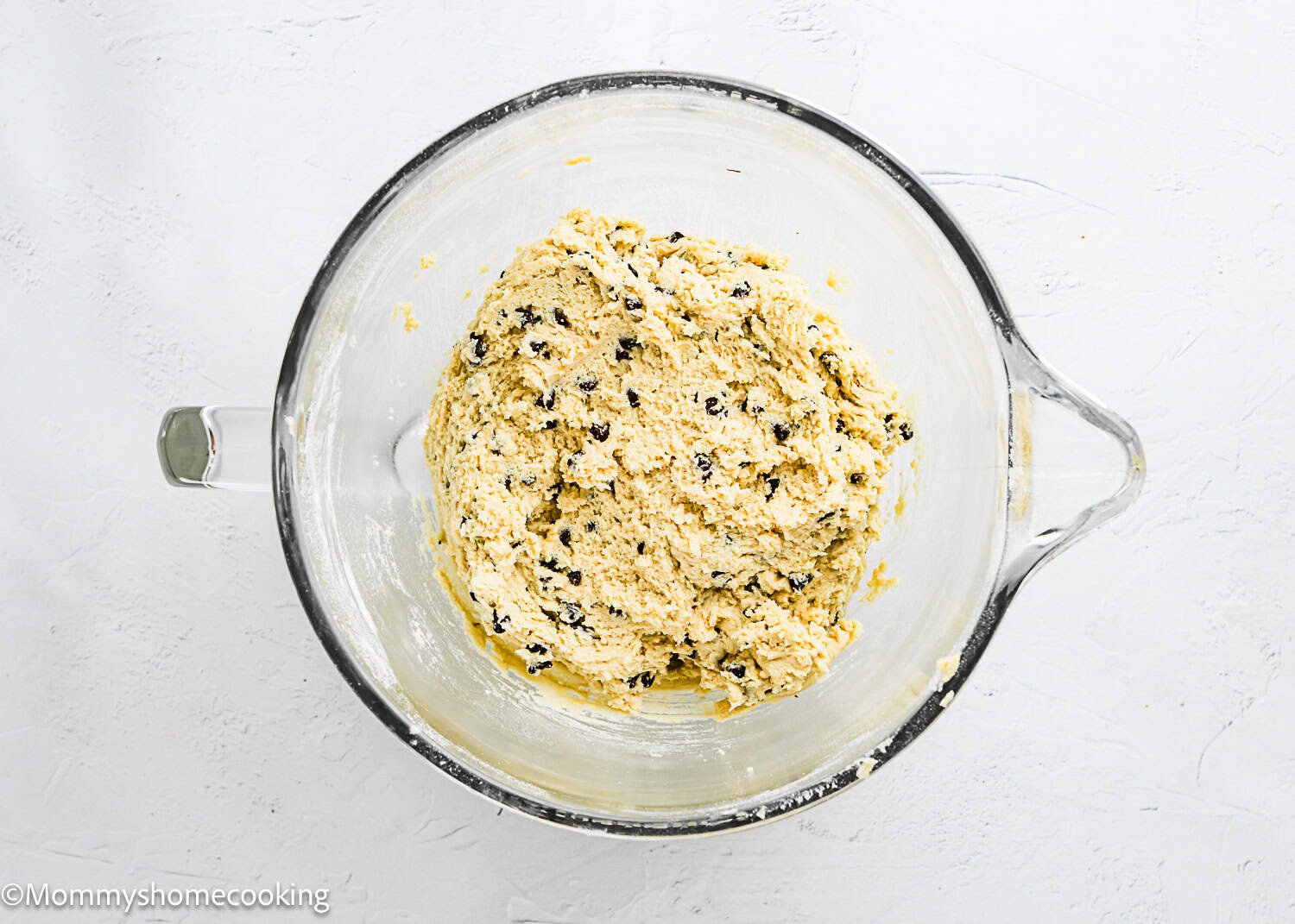 A glass mixing bowl containing dough for eggless chocolate chip cookie bars sits on a white surface.