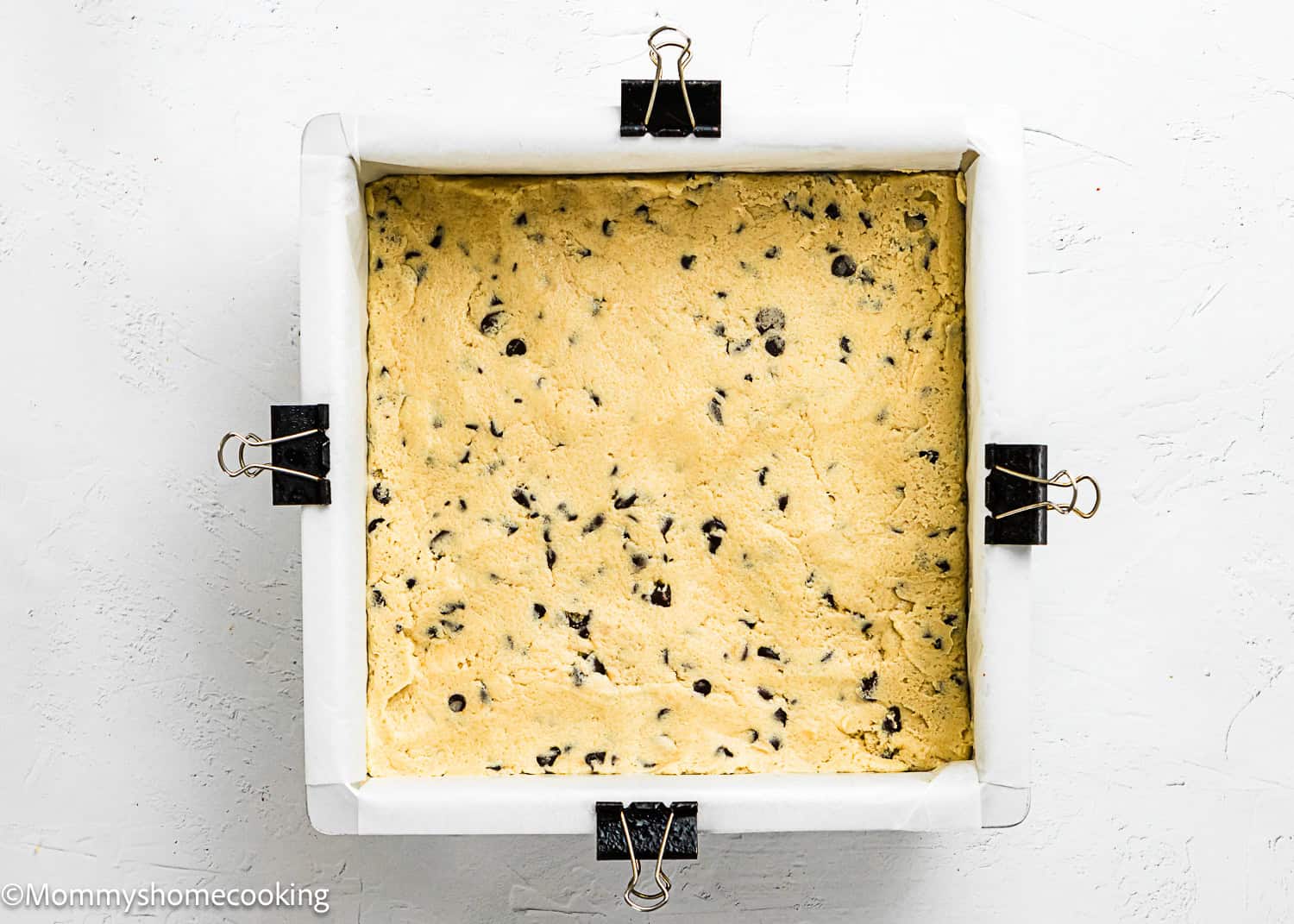 Square baking pan lined with parchment paper, filled with Eggless Chocolate Chip Cookie Bars dough, secured at the sides with binder clips.
