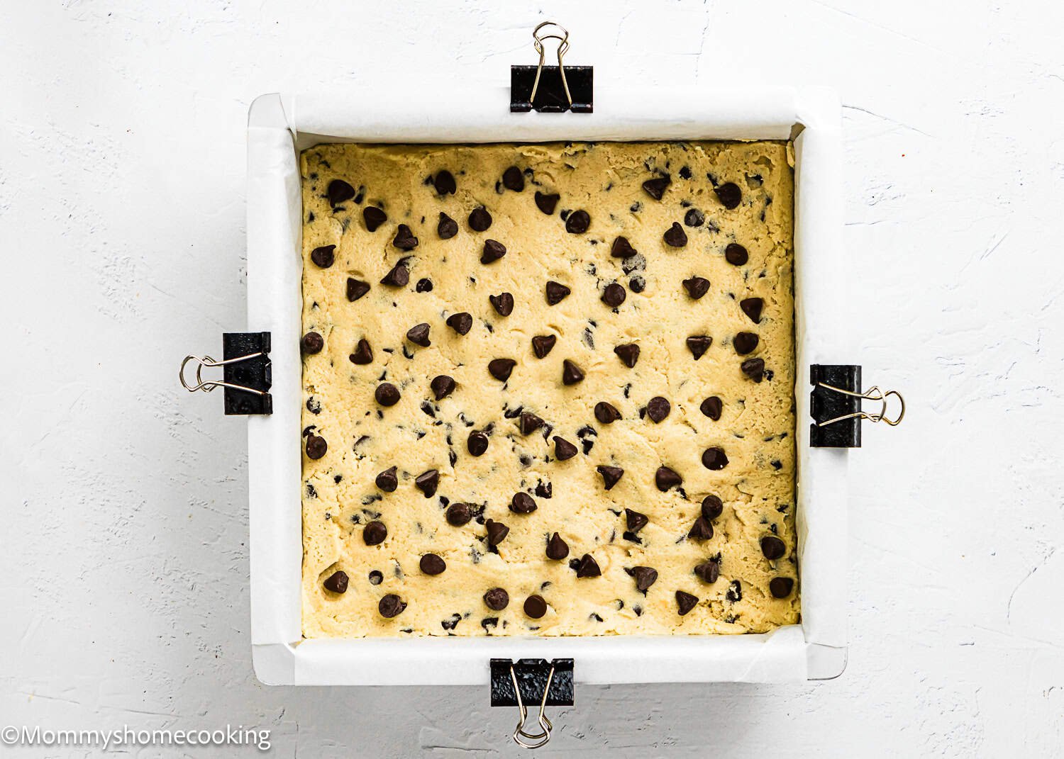 Square pan lined with parchment paper and secured with clips, filled with unbaked Eggless Chocolate Chip Cookie Bars dough, viewed from above.