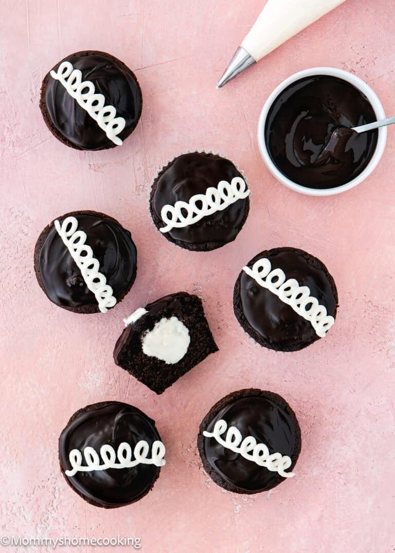 Seven Eggless Chocolate Cream Cupcakes (hostess cupcakes) with glossy tops and white icing swirls are arranged on a pink surface; one is cut open to reveal a creamy filling. A piping bag and bowl of chocolate are nearby.