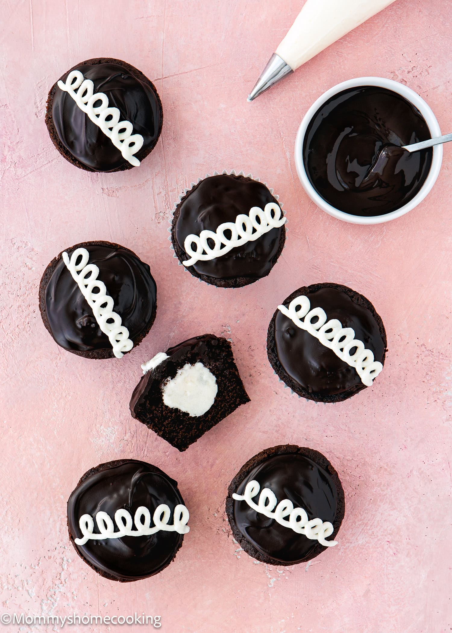 Seven Eggless Chocolate Cream Cupcakes (hostess cupcakes) with glossy tops and white icing swirls are arranged on a pink surface; one is cut open to reveal a creamy filling. A piping bag and bowl of chocolate are nearby.