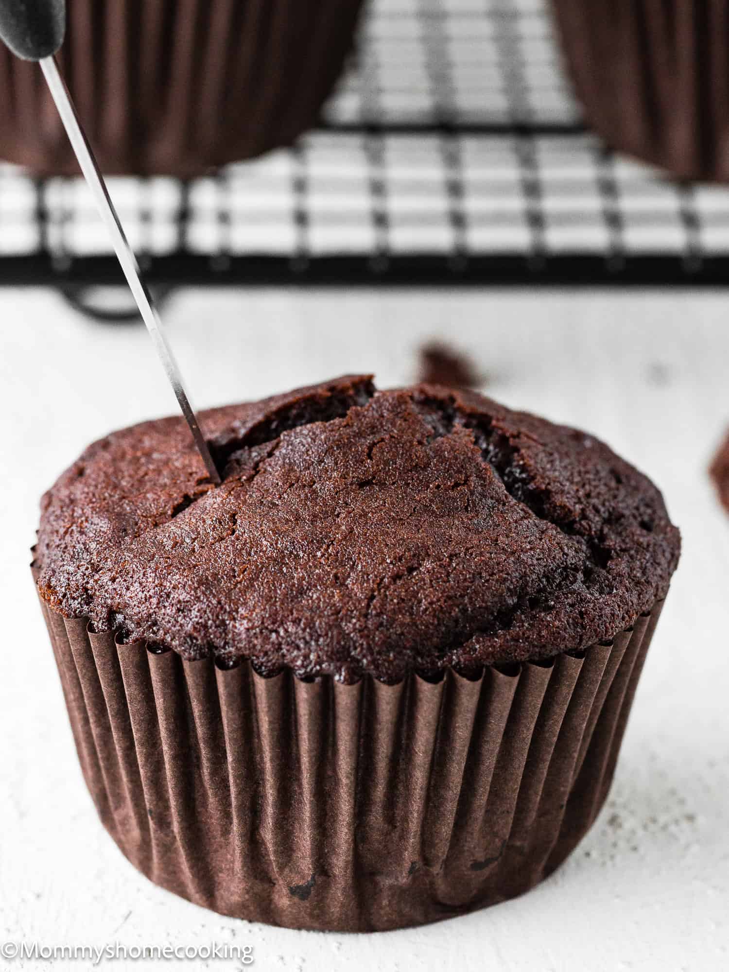 An Eggless Chocolate Cream Cupcake (hostess cupcake) in a brown paper liner is being cut. A cooling rack with more cupcakes is visible in the background.