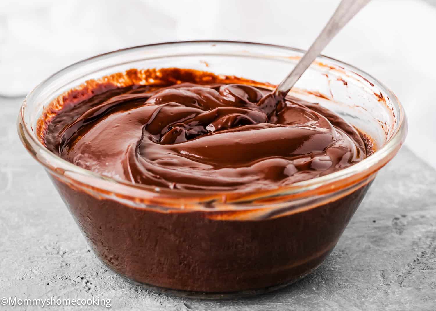 A clear glass bowl filled with smooth, glossy chocolate ganache for Eggless Chocolate Cream Cupcakes (hostess cupcakes), with a metal spoon resting inside.