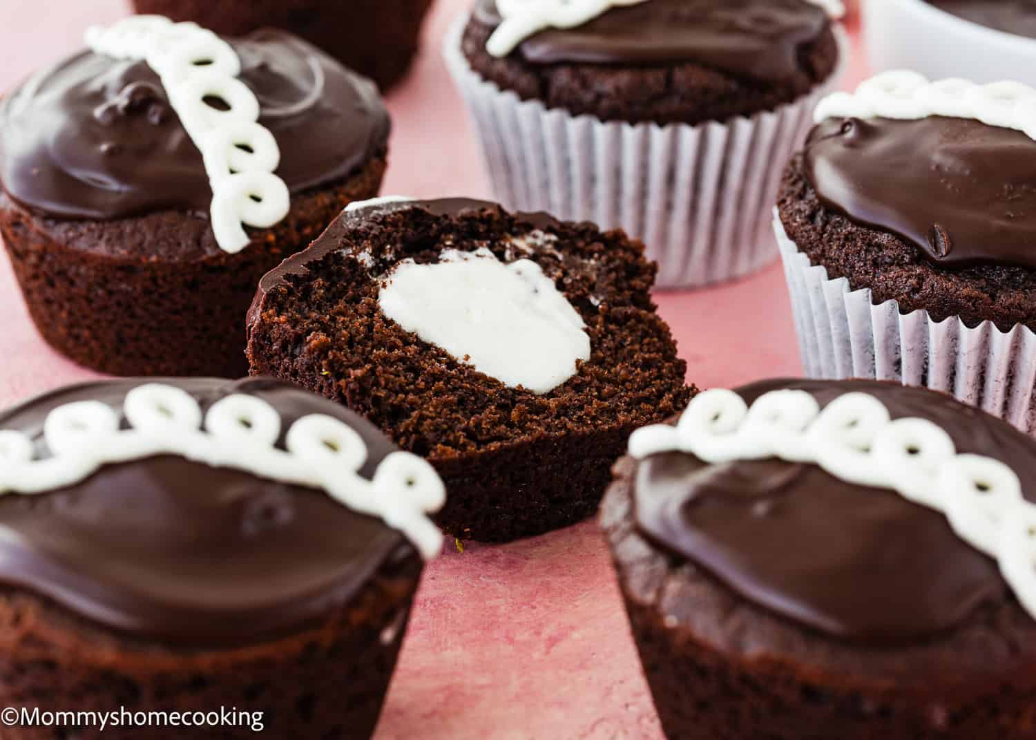 Eggless Chocolate Cream Cupcakes (hostess cupcakes) with chocolate frosting and white icing decoration; one cupcake is cut in half to reveal a creamy filling inside.