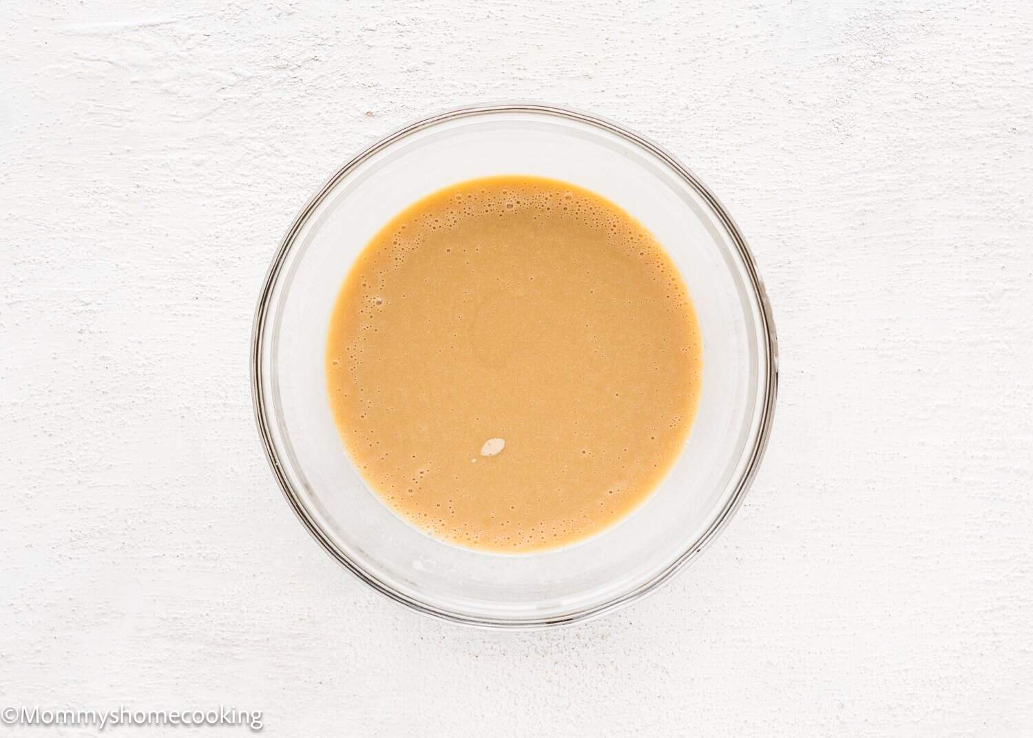 A clear glass bowl containing wet ingredientes  for Eggless Chocolate Cream Cupcakes (hostess cupcakes) sits on a white surface.