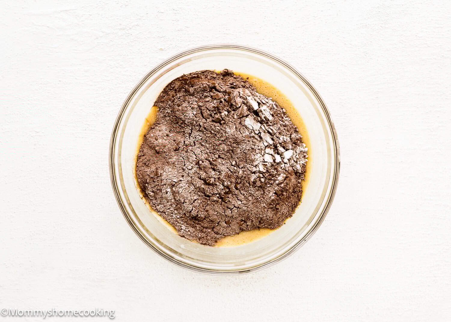 A clear glass bowl containing wet and dry ingredientes  for Eggless Chocolate Cream Cupcakes (hostess cupcakes) sits on a white surface.