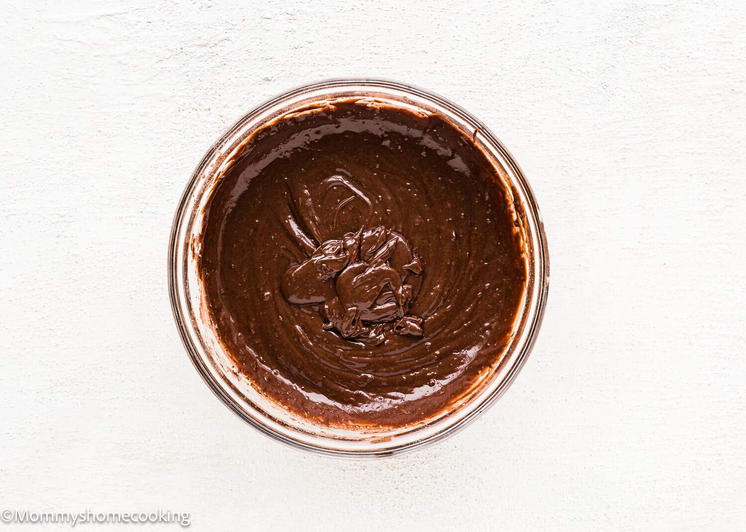 A glass bowl filled with smooth, mixed chocolate batter for Eggless Chocolate Cream Cupcakes (hostess cupcakes) sits on a white textured surface.