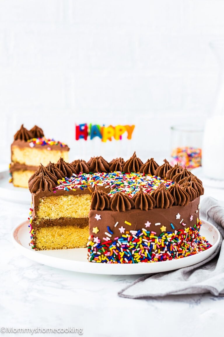 An Eggless Classic Yellow Birthday Cake with chocolate frosting and colorful sprinkles sits on a plate, one slice removed. "Happy" candles and a glass of milk are in the background.