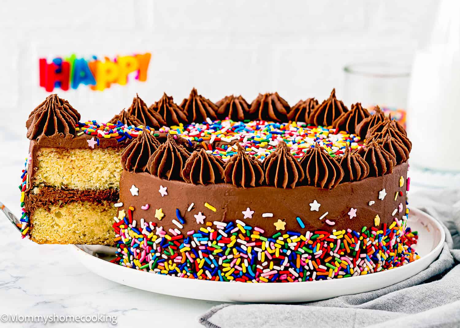 A round Eggless Classic Yellow Birthday Cake with chocolate frosting and colorful sprinkles, one slice being removed; "Happy" sign in the background.