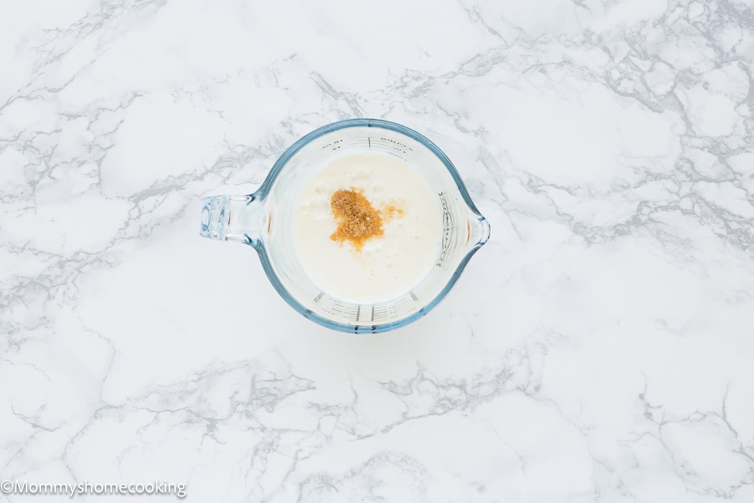 A glass measuring cup filled with milk and a small amount of brown sugar sits on a white marble surface, perfect for making an Eggless Classic Yellow Birthday Cake.
