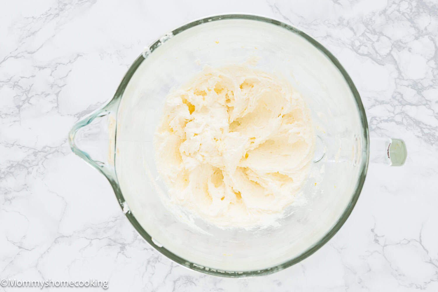 A glass mixing bowl filled with a creamed butter and sugar mixture for an Eggless Classic Yellow Birthday Cake sits on a marble countertop.