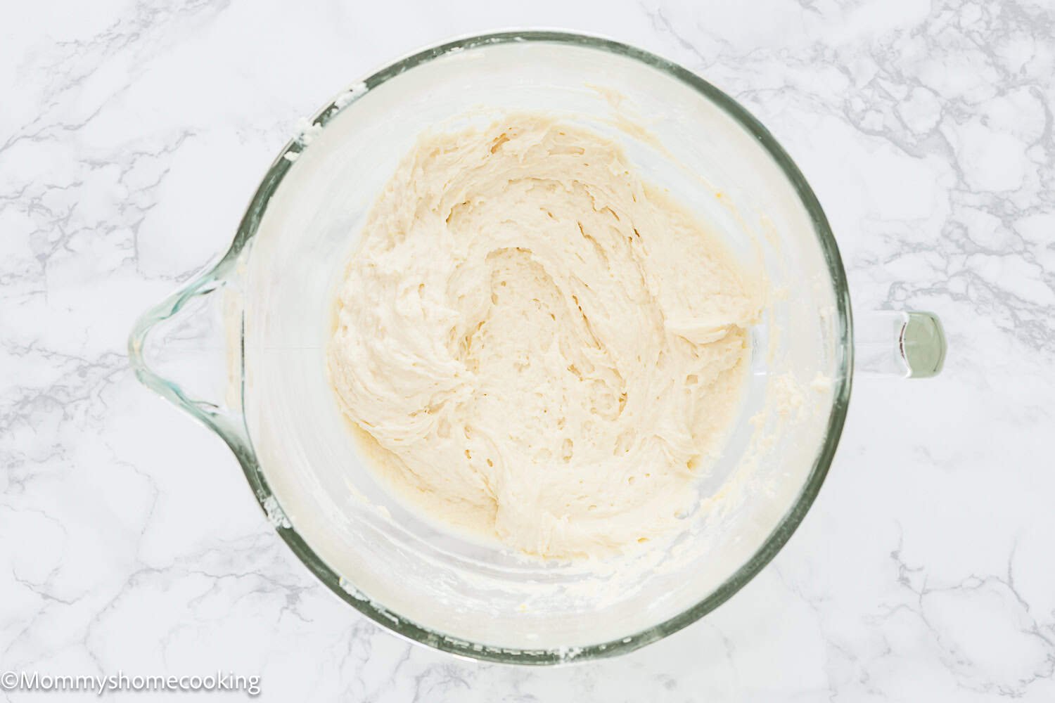 A glass mixing bowl containing thick, creamy Eggless Classic Yellow Birthday Cake batter sits on a white marble surface.