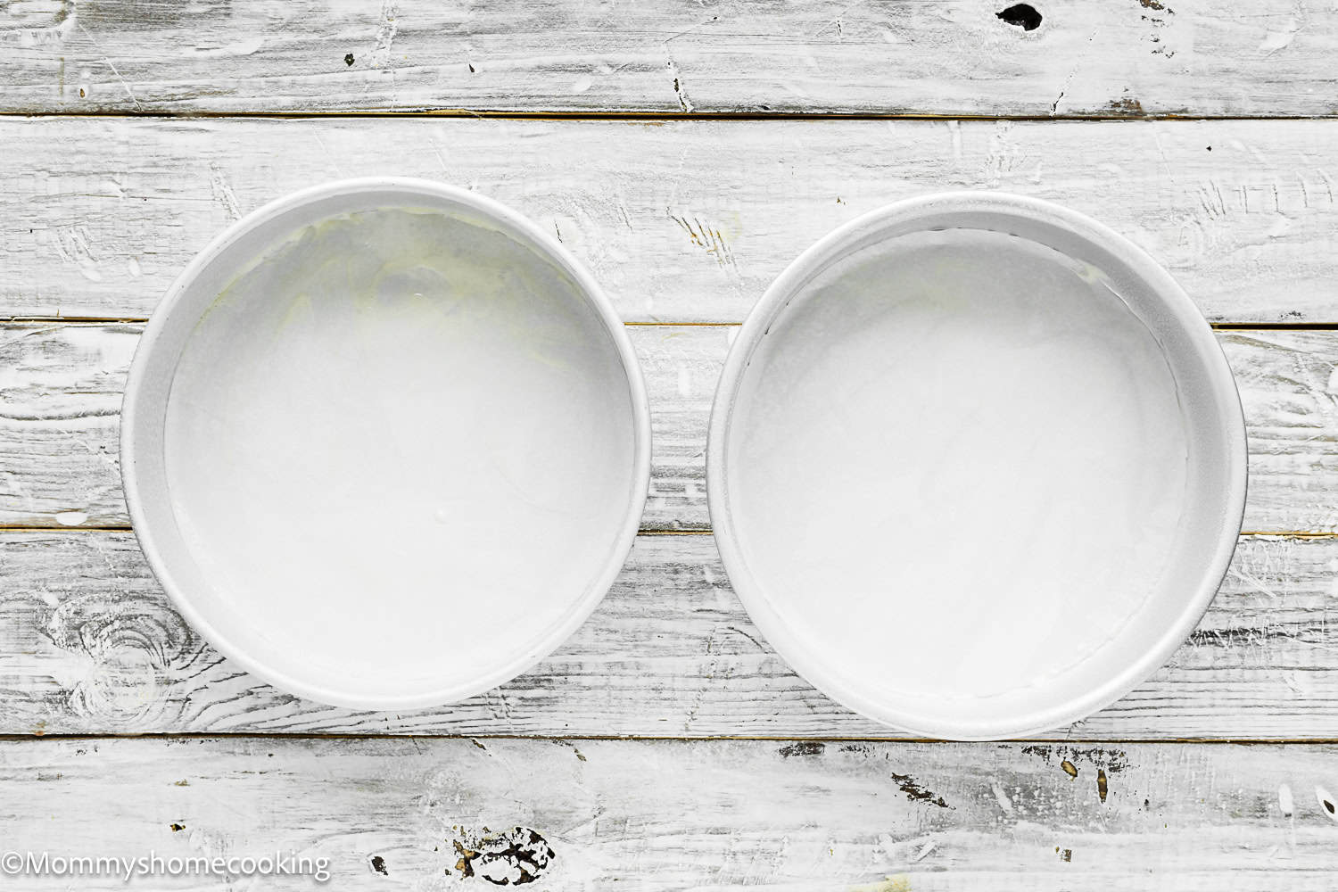 Two round cake pans coated with flour and grease are placed side by side on a white wooden surface, ready to bake an Eggless Classic Yellow Birthday Cake.