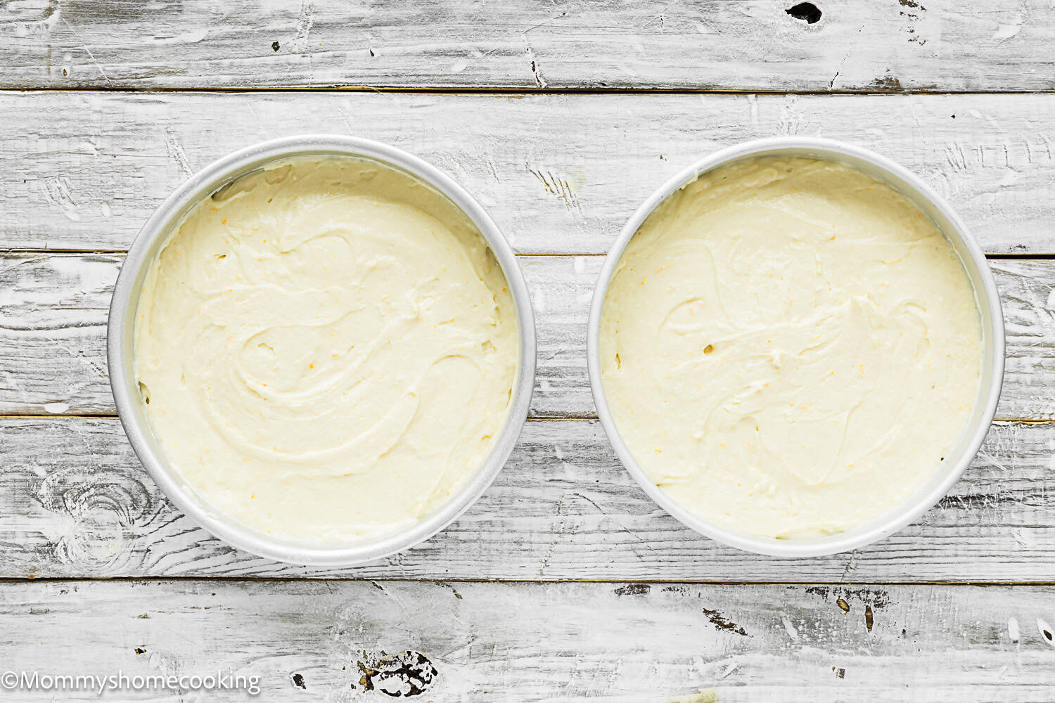 Two round cake pans filled with eggless classic yellow birthday cake batter sit on a white wooden surface.