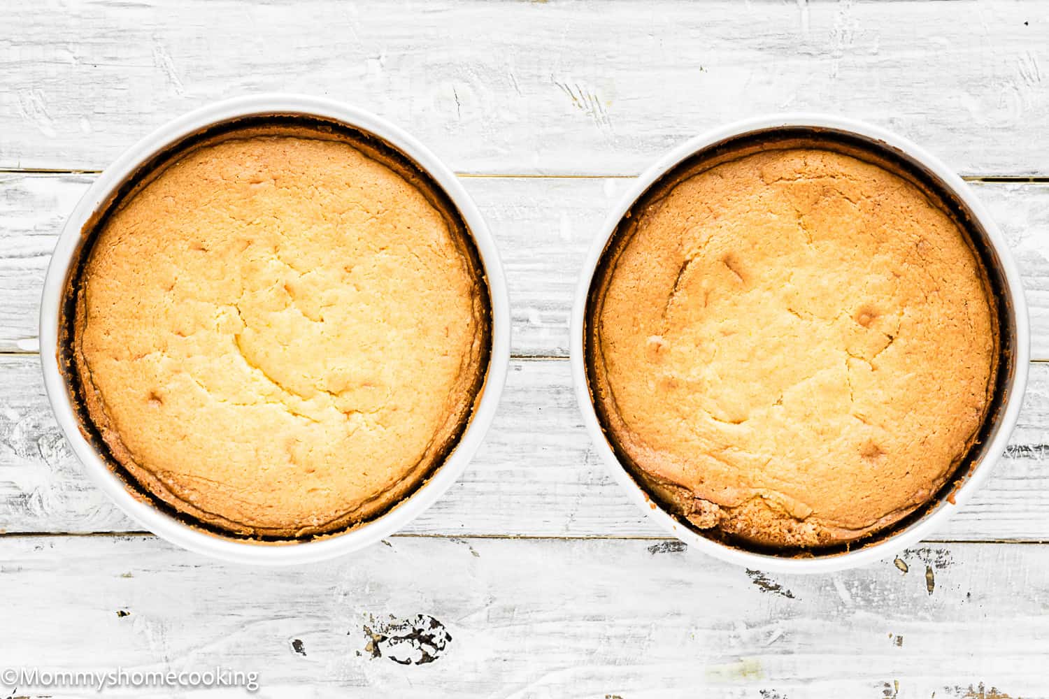 Two round, golden-brown Eggless Classic Yellow Birthday Cakes in white baking dishes sit side by side on a white wooden surface.