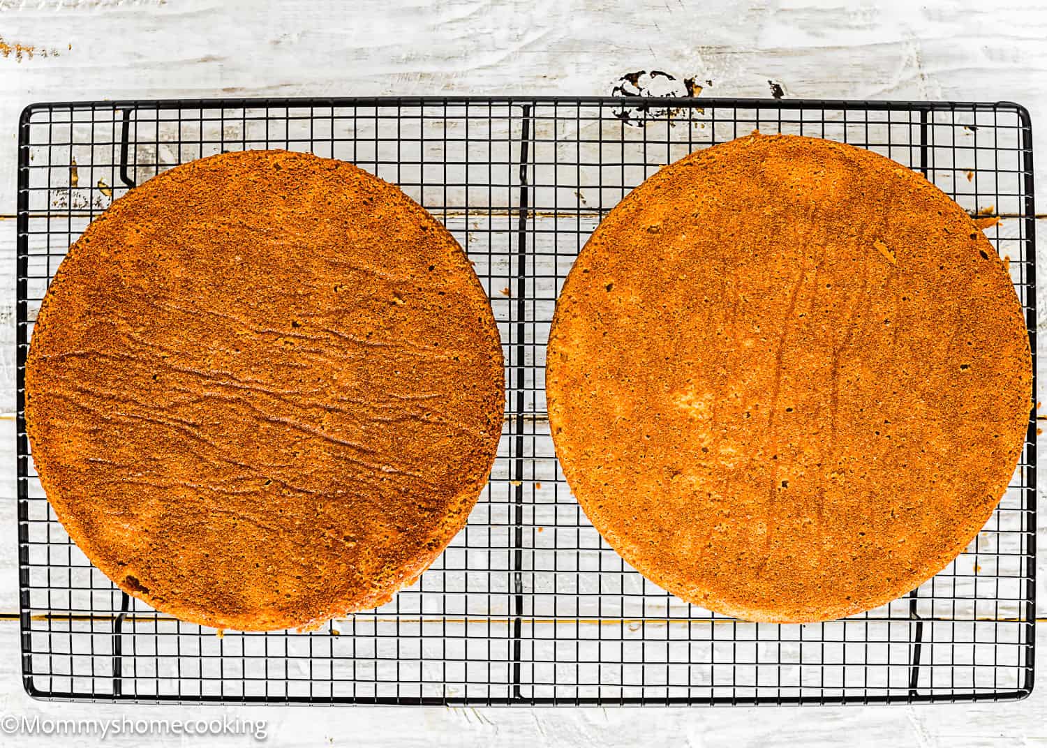 Two round, golden-brown Eggless Classic Yellow Birthday Cake layers are cooling on a black wire rack set on a white surface.