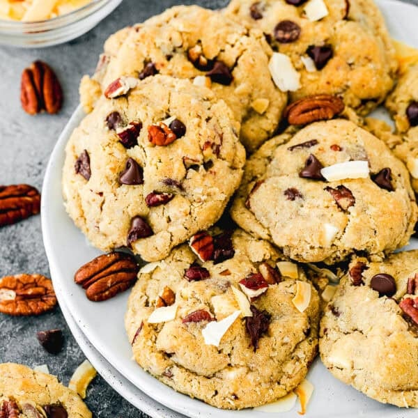 A plate of Eggless Cowboy Cookies topped with chopped pecans and toasted coconut flakes, with extra pecans, chocolate chips, and coconut flakes scattered nearby.