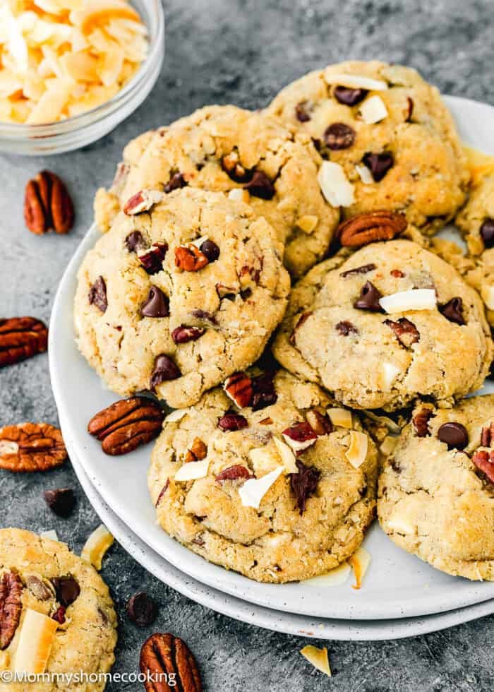 A plate of Eggless Cowboy Cookies topped with chopped pecans and toasted coconut flakes, with extra pecans, chocolate chips, and coconut flakes scattered nearby.