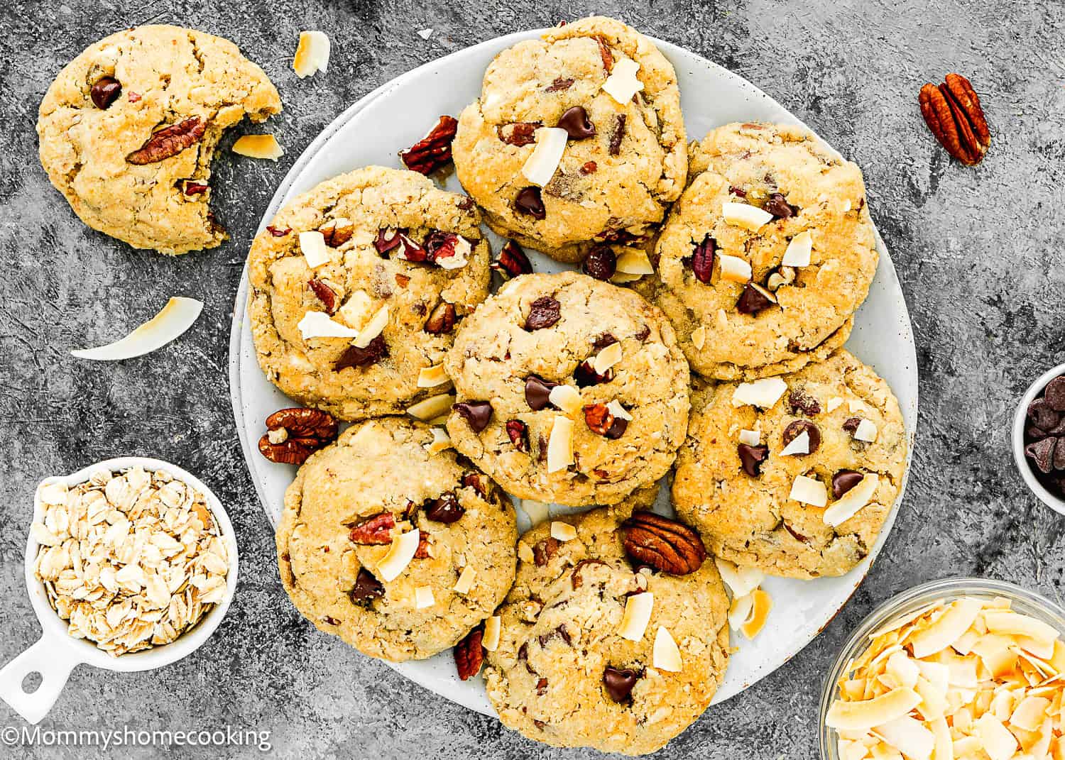 A plate of Eggless Cowboy Cookies with pecans and coconut flakes, surrounded by a bowl of oats, pecans, and coconut flakes on a gray surface.