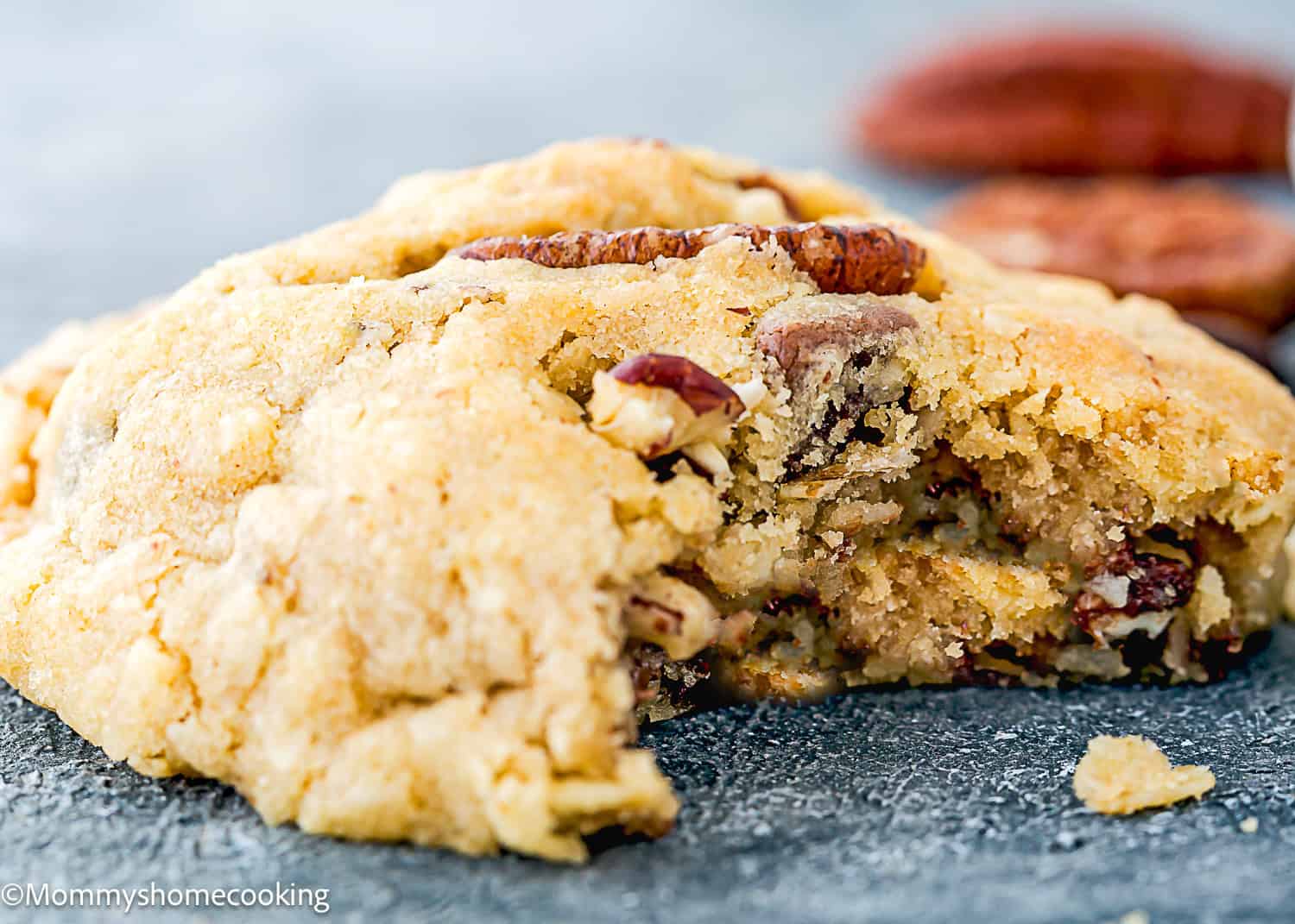 A close-up of a thick, crumbly Eggless Cowboy Cookie with pecans, partially eaten, resting on a gray surface.