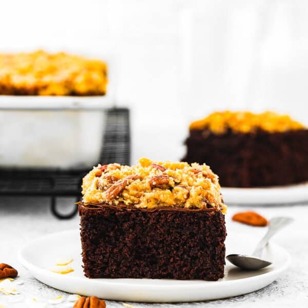 A slice of Eggless German Chocolate Sheet Cake topped with coconut pecan frosting sits on a white plate with a spoon; more cake and pecans are in the background.