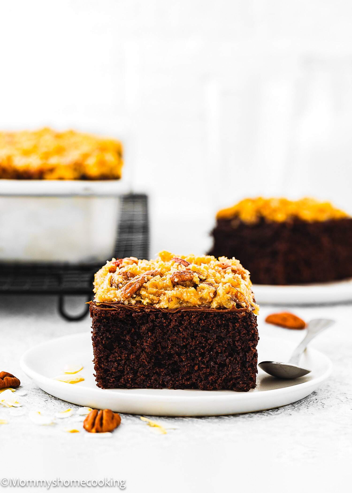 A slice of Eggless German Chocolate Sheet Cake topped with coconut pecan frosting sits on a white plate with a spoon; more cake and pecans are in the background.