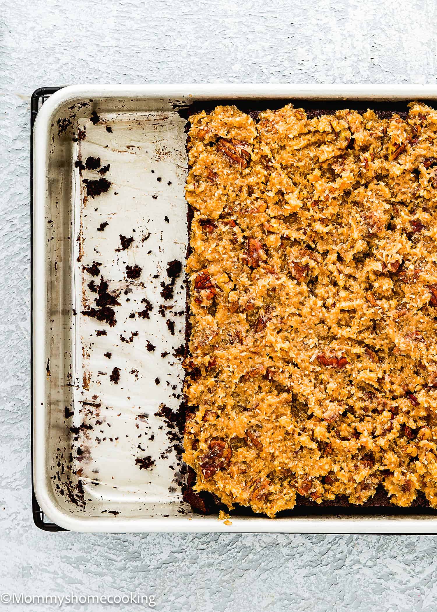 A partially filled baking pan with a textured, crumbly dessert mixture covering about two-thirds of the surface, featuring Eggless German Chocolate Sheet Cake.