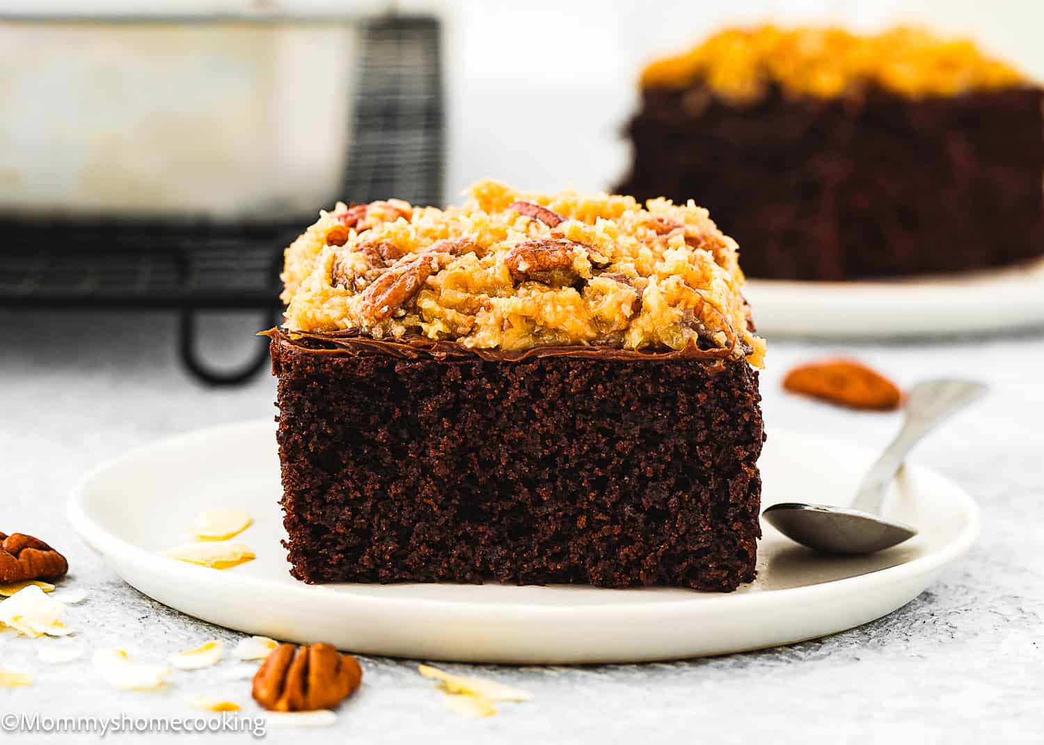 A slice of Eggless German Chocolate Sheet Cake with coconut-pecan frosting sits on a white plate with a spoon, pecans, and coconut flakes nearby. Another slice is blurred in the background.