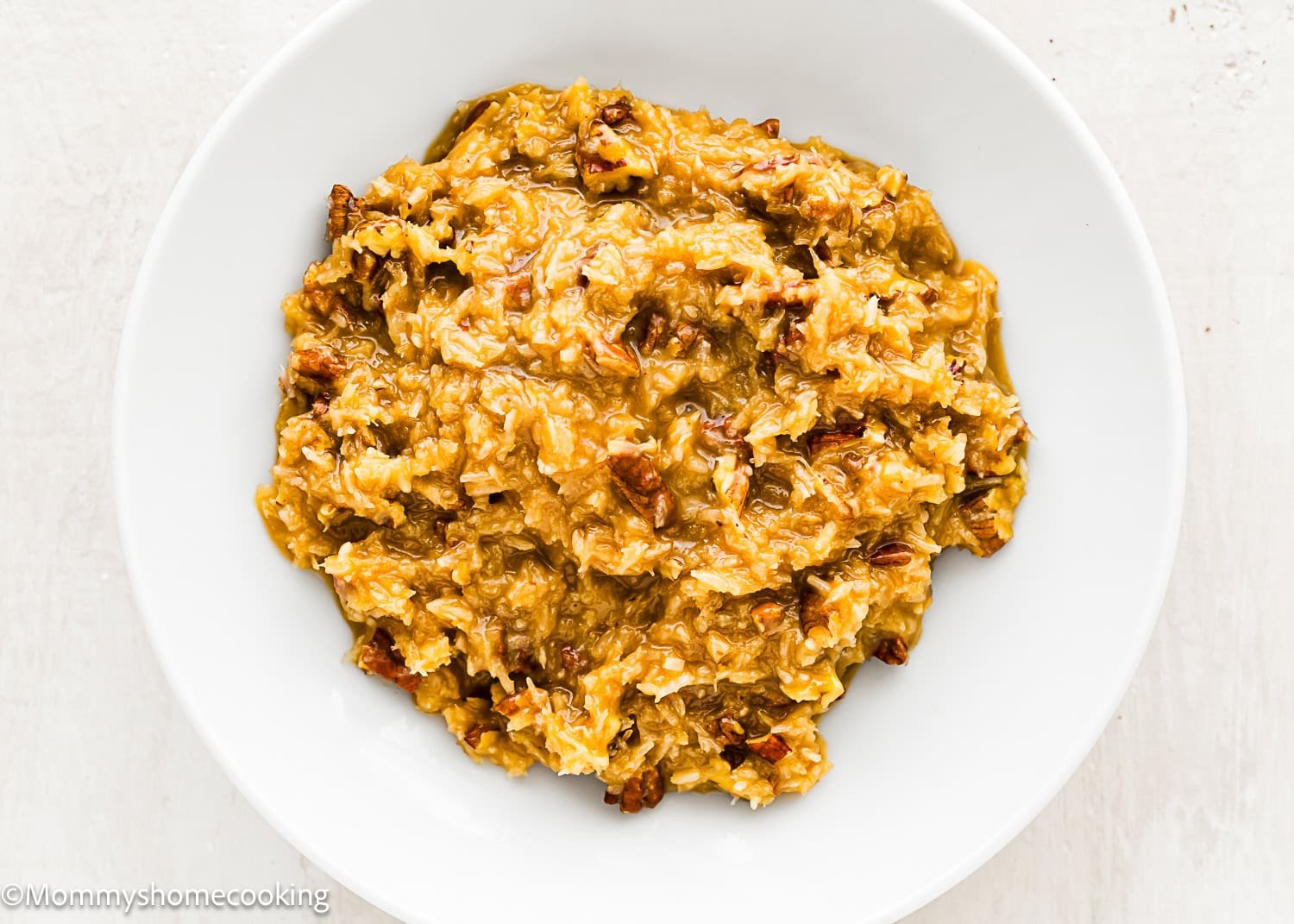 A white plate holds a serving of moist, golden brown coconut and pecan frosting with visible pecan pieces, perfect for topping an Eggless German Chocolate Sheet Cake.
