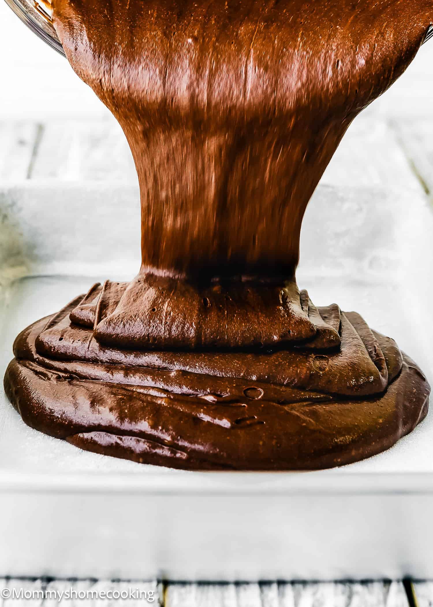 Thick chocolate brownie batter for an Eggless German Chocolate Sheet Cake is being poured from a bowl into a greased square baking pan.