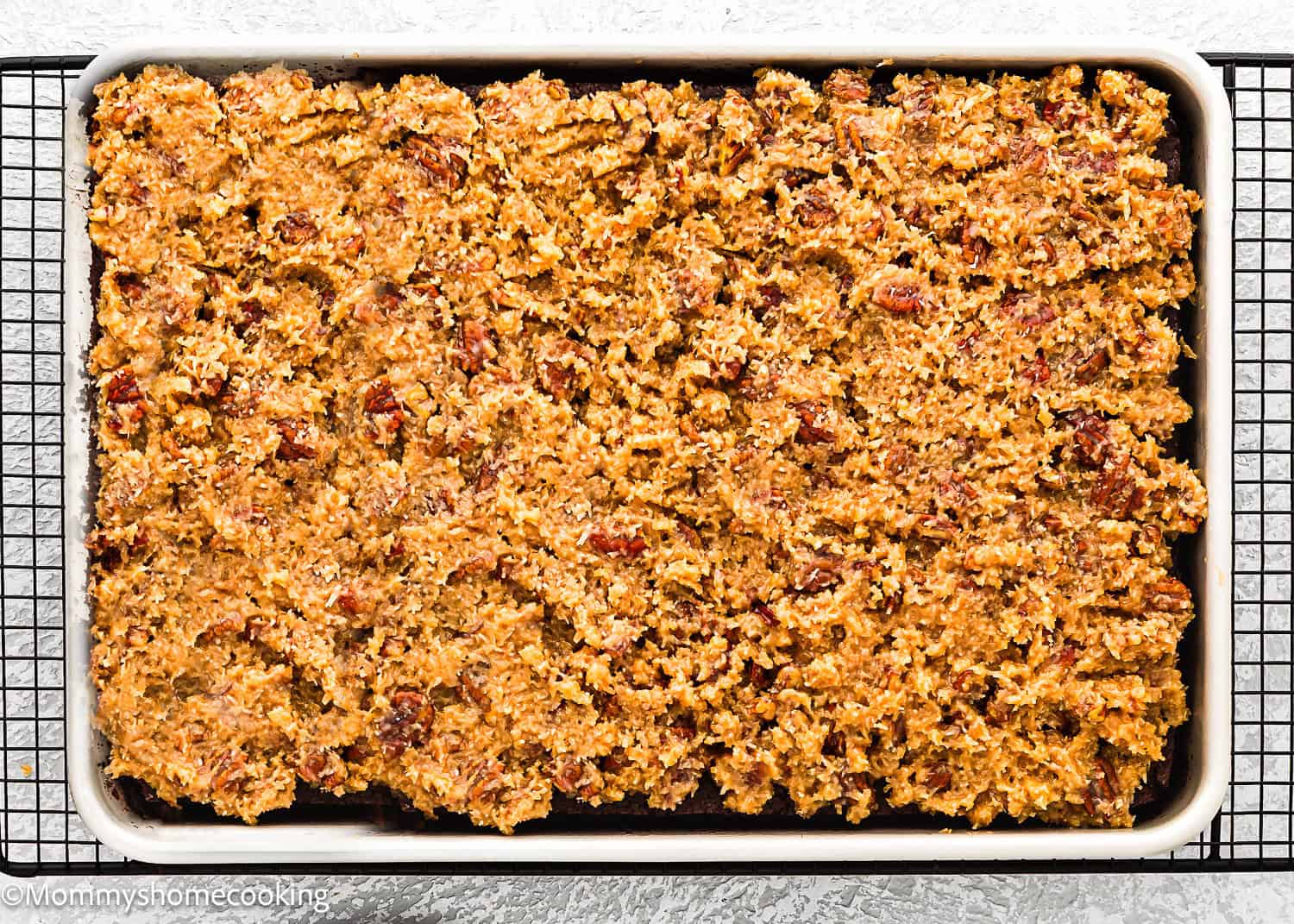 A rectangular baking pan filled with an Eggless German Chocolate Sheet Cake, topped with a textured, golden-brown coconut and pecan mixture, cooling on a wire rack.