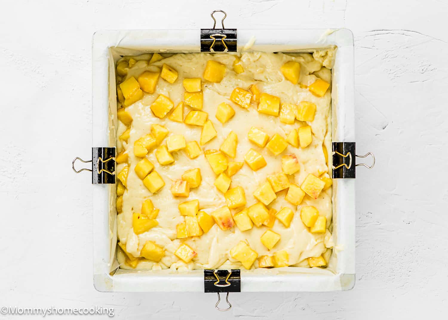 A square baking pan lined with parchment paper and clipped at the sides, filled with Eggless Peach Coffee Cake batter and topped with diced peach pieces.