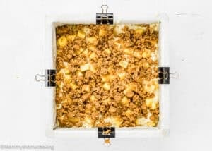 A square baking pan lined with parchment paper and secured with binder clips, filled with an unbaked eggless peach coffee cake crumble mixture.