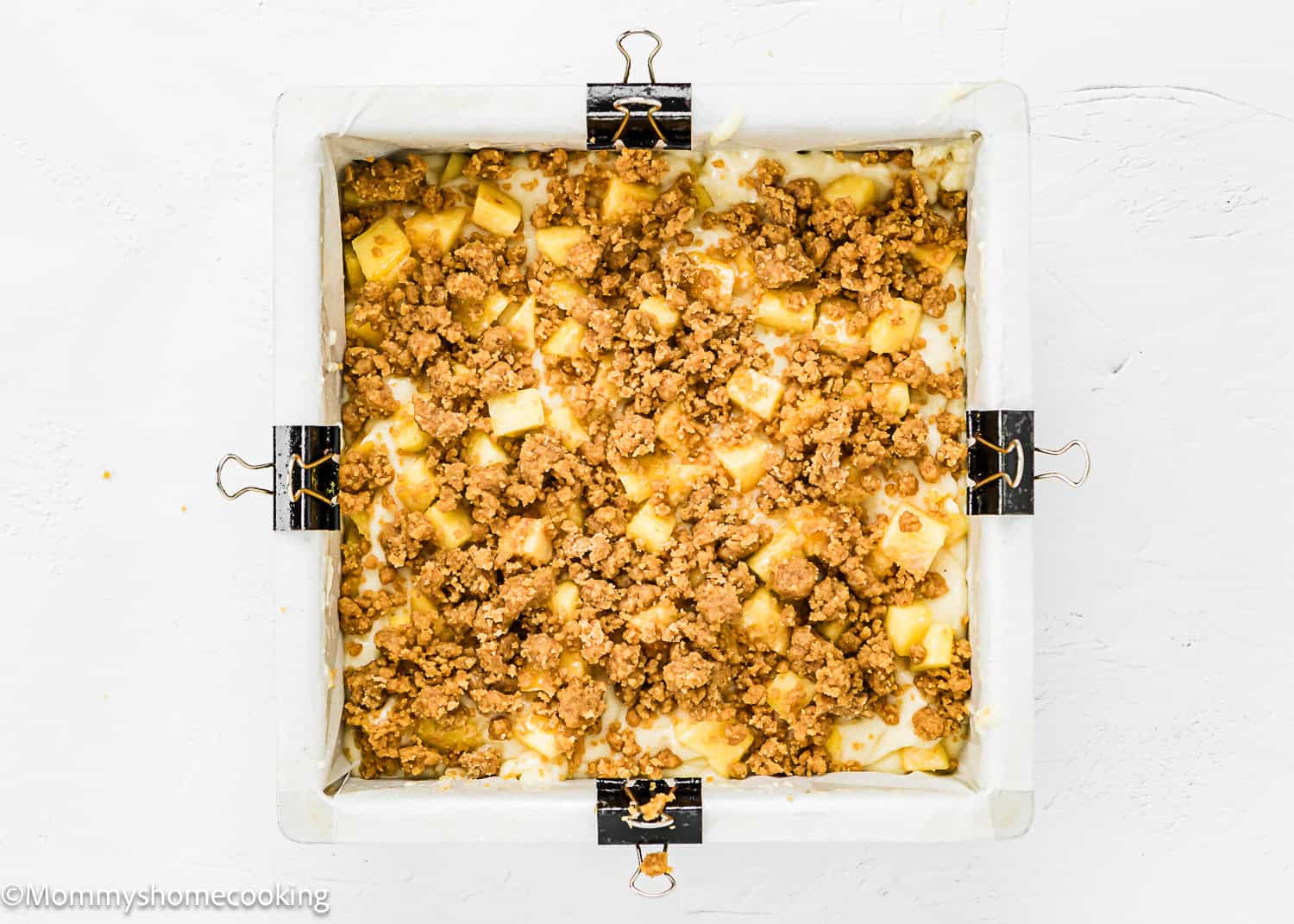 A square baking pan lined with parchment paper and secured with binder clips, filled with an unbaked eggless peach coffee cake batter and crumble mixture.