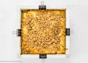 A square baking pan lined with parchment paper holds an Eggless Peach Coffee Cake topped with a golden brown streusel.
