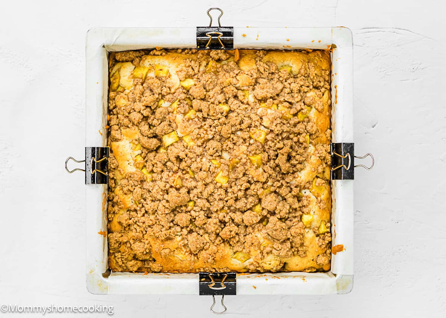 A square baking pan lined with parchment paper holds an Eggless Peach Coffee Cake topped with a golden brown streusel.