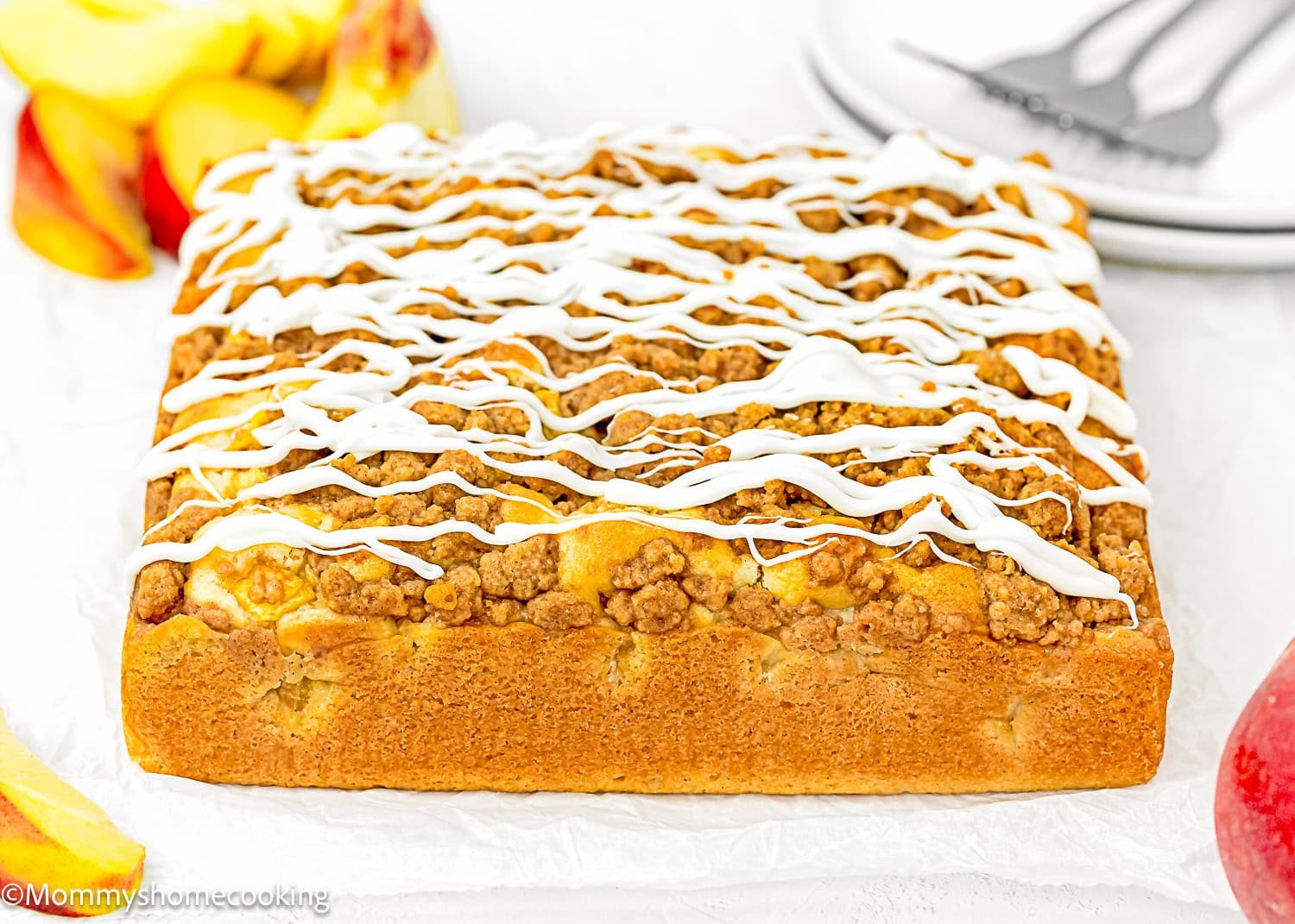 A square Eggless Peach Coffee Cake with a crumb topping and white icing drizzle is displayed on parchment paper, with apple slices and plates in the background.