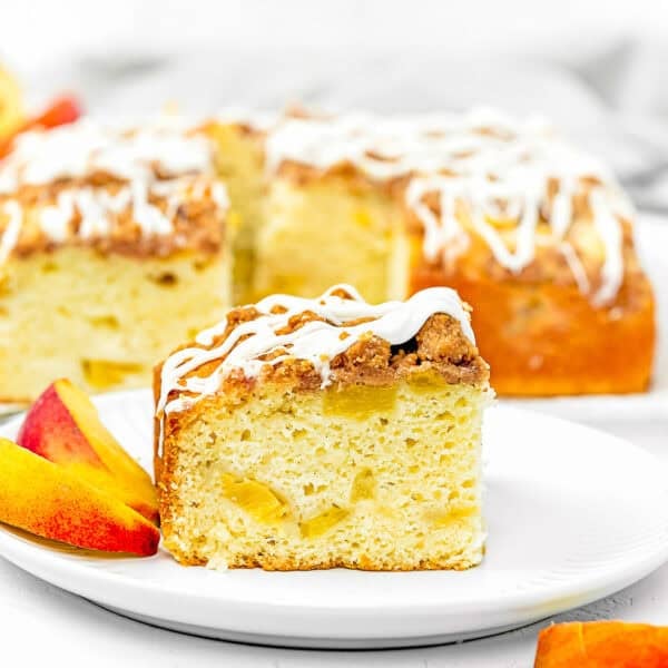 A slice of Eggless Peach Coffee Cake with crumb topping and visible fruit pieces sits on a white plate, alongside peach slices and more cake in the background.