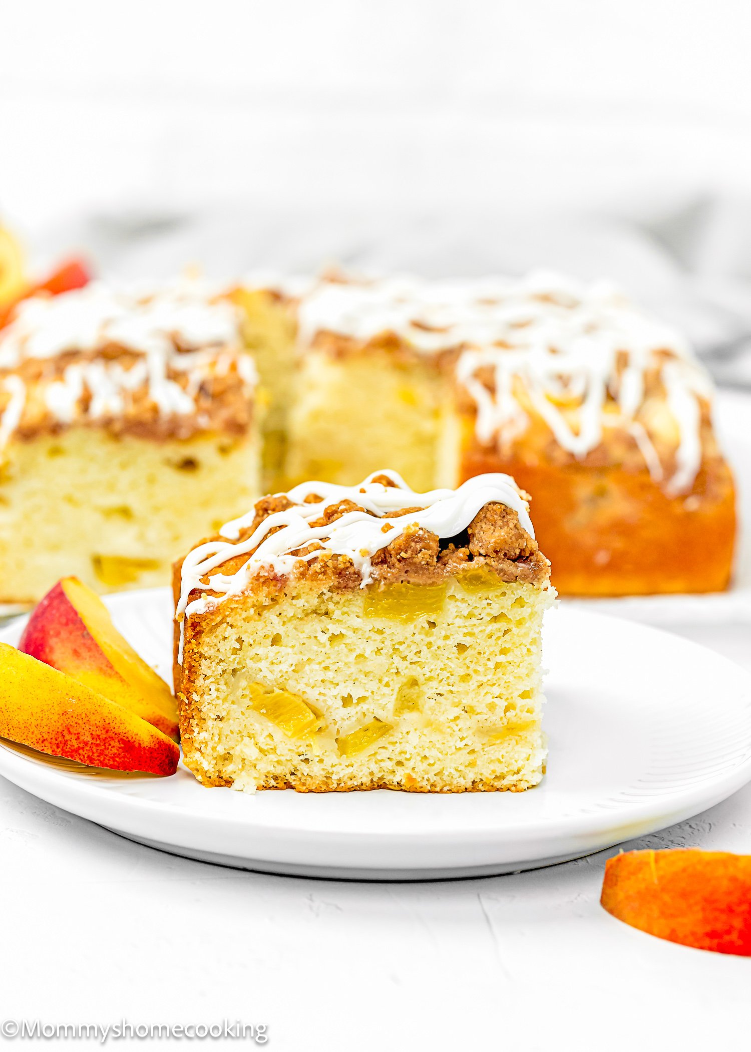 A slice of Eggless Peach Coffee Cake with crumb topping and visible fruit pieces sits on a white plate, alongside peach slices and more cake in the background.